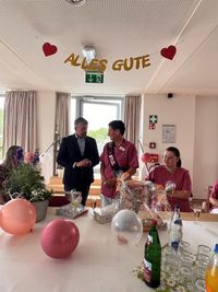 Das Bild zeigt 4 Personen in einem Raum. Zwei von ihnen stehen: Ein Mann im Anzug und eine Frau im rosafarbenen Kasack. Sie trägt eine "Ruhestand"-Schärpe.