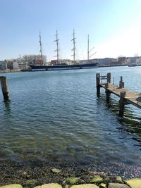 Blick über einen Fluss. Am anderen Ufer liegt ein großes Segelschiff mit vier Masten.