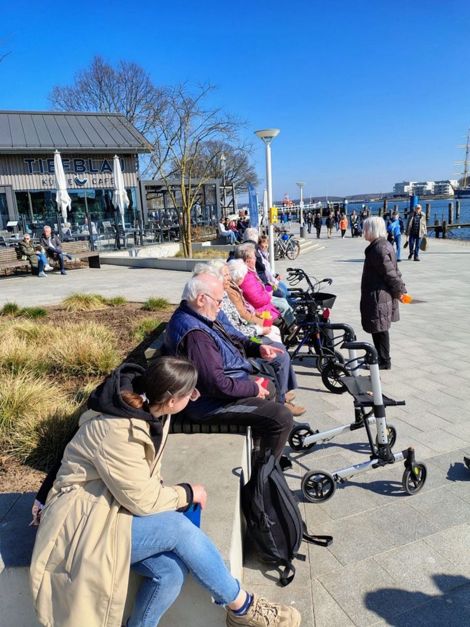 Eine Gruppe Senior*innen sitzt auf einer Bank in der Sonne. Vor ihnen stehen diverse Rollatoren.