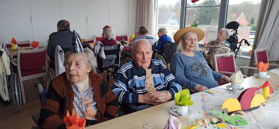 Neun u.a. mit Hüten verkleidete Senior*innen sitzen an bunt geschmückten Kaffeetafeln in einem großen Saal.